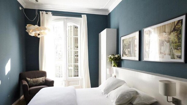 A bedroom with a large bed, a chair, a window with curtains, and framed pictures on the wall. - Bed and Break Barcelona (Barcelona)
