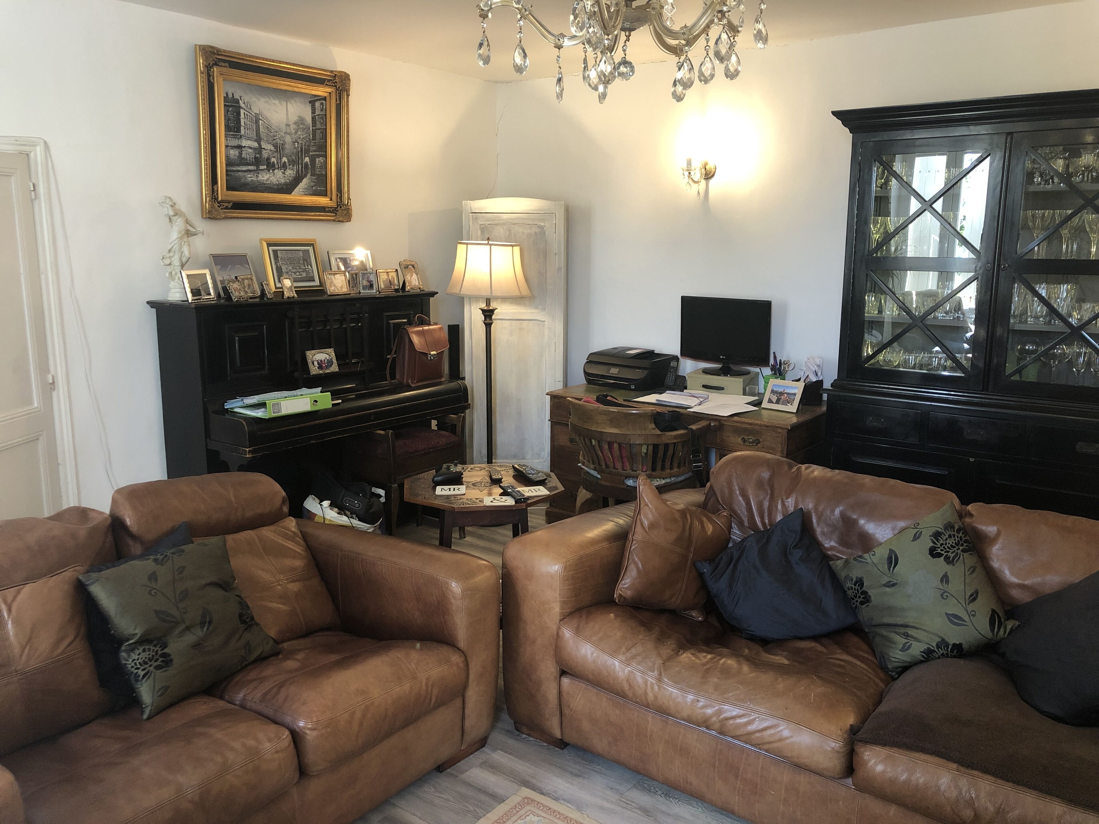 A living room with two brown leather sofas, a piano, a desk with a computer, and a chandelier.