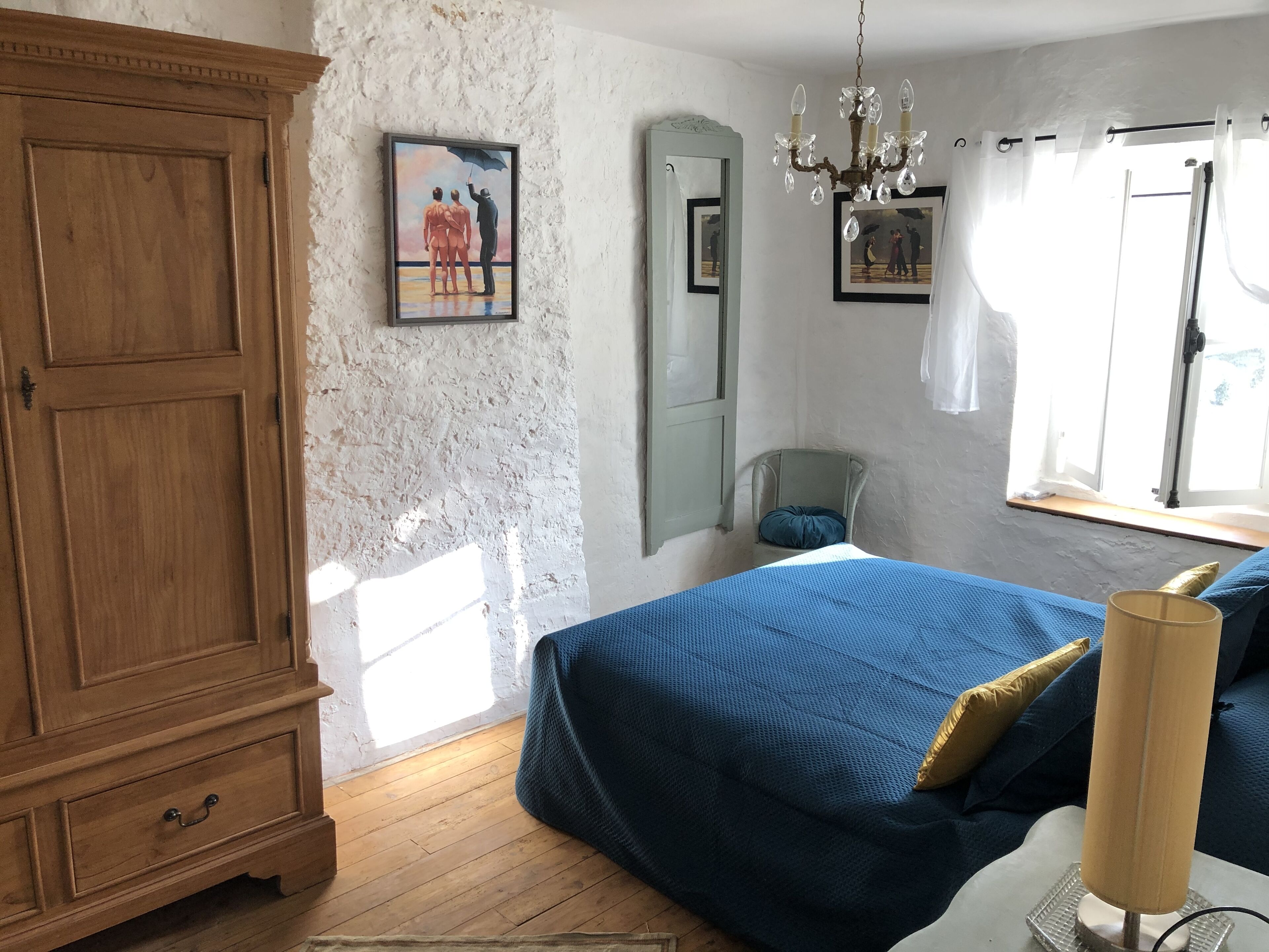 Un dormitorio con un armario de madera, una cama con cubrecama azul, una araña, un espejo y una ventana con cortinas.