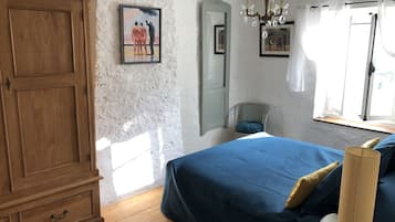 Un dormitorio con un armario de madera, una cama con cubrecama azul, una araña, un espejo y una ventana con cortinas.