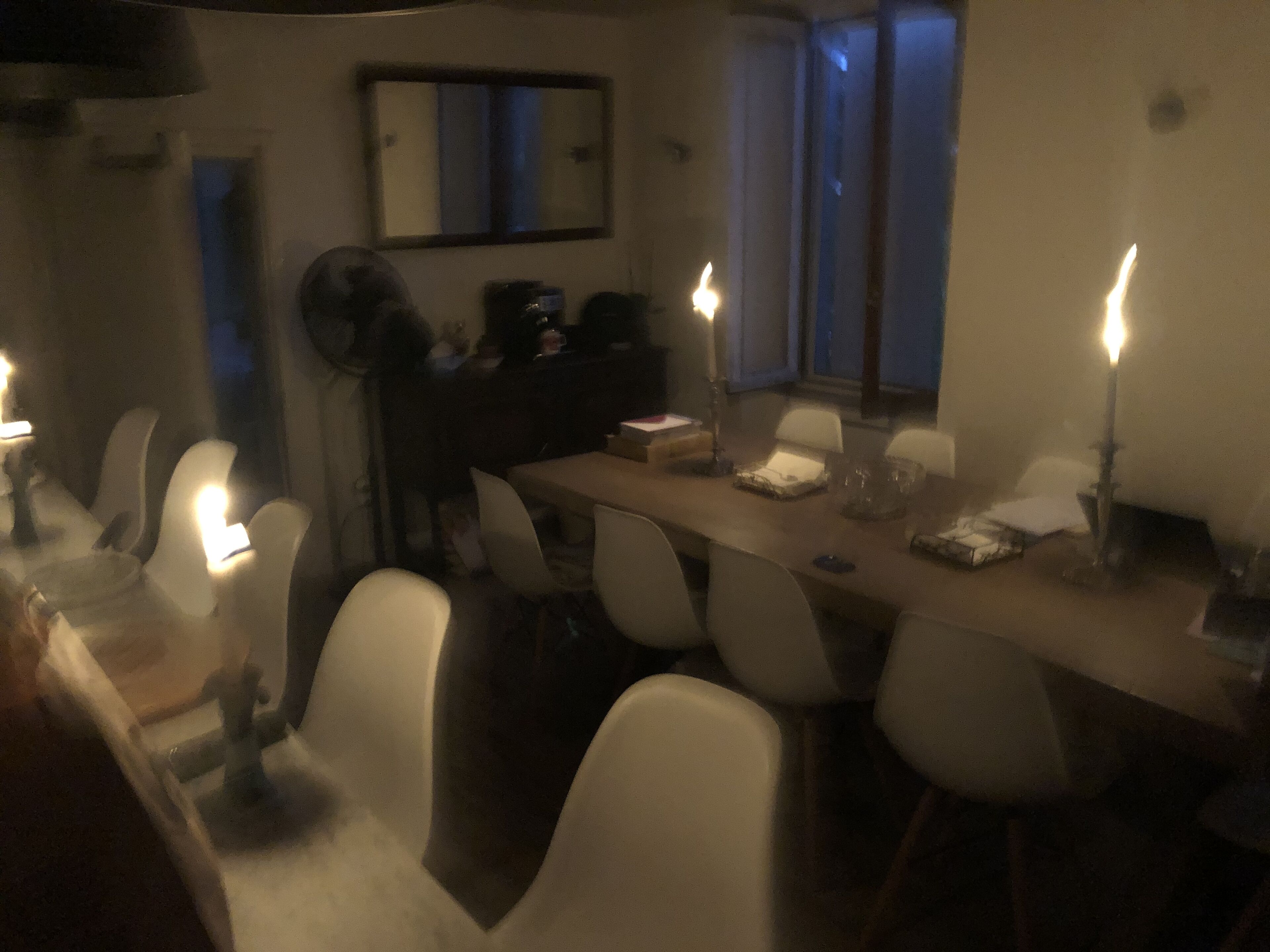 A dining area with a long table, white chairs, and candles. There is a mirror on the wall and a window with a view of the outside.