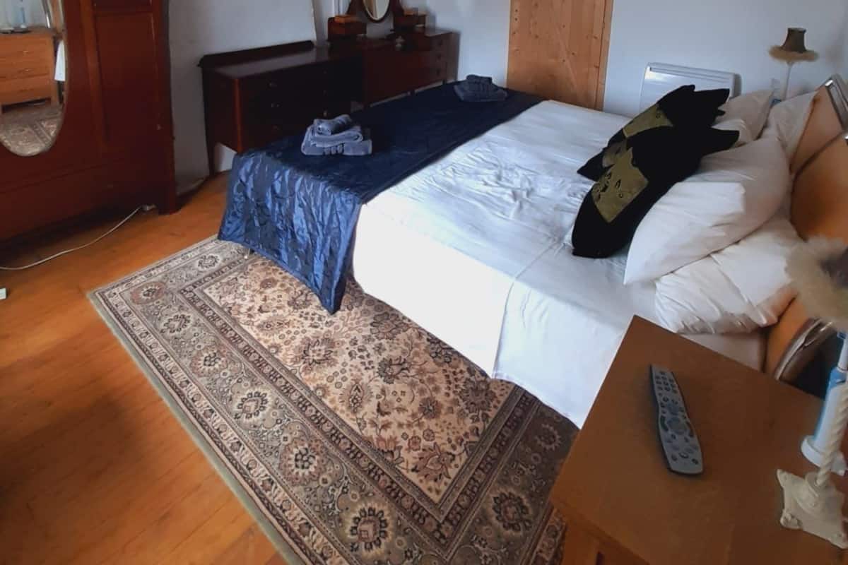 A bedroom with a wooden ceiling, a bed with white bedding, a blue bedspread, a wooden wardrobe, a mirror, a chandelier, and a patterned rug.