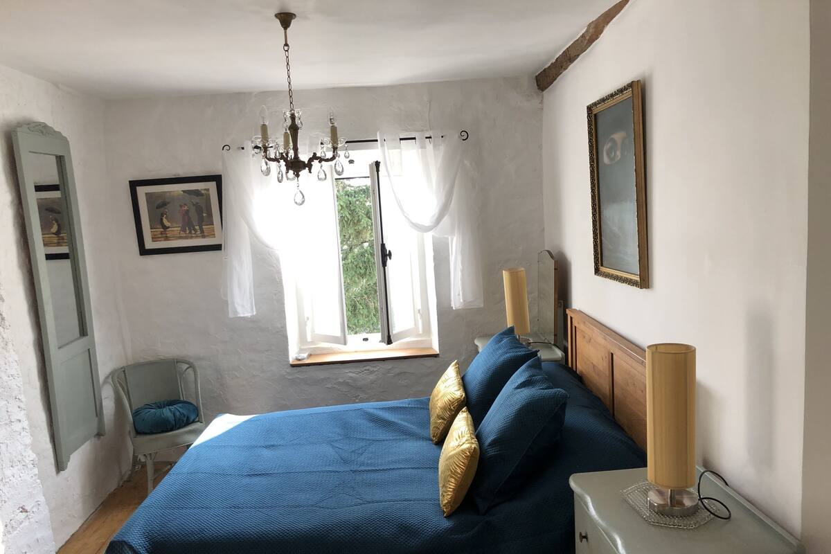 A bedroom with a bed, a nightstand, a lamp, a window with curtains, a mirror, and a painting on the wall.