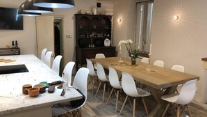 Un comedor con una mesa larga, sillas blancas y mantel de madera. Se observa una zona de cocina con estufa y armarios, además de un televisor montado en la pared.