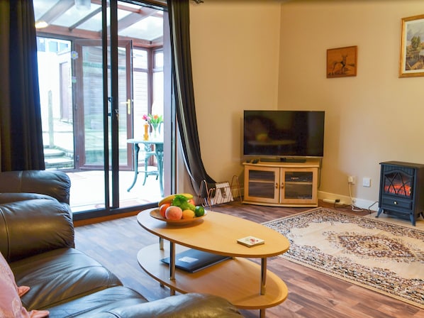 A living room with a leather sofa, a small round table with a fruit bowl, a flat-screen TV on a stand, and a wood-burning stove.