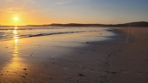 Une plage au coucher du soleil, avec des vagues calmes, un ciel dégagé et des collines lointaines.