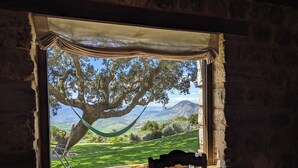Une pièce rustique dotée d’une grande fenêtre donnant sur un paysage luxuriant, d’un hamac et d’une table avec une chaise.