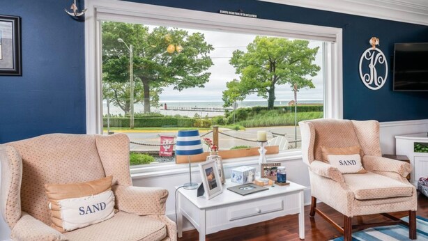 Une pièce comprenant deux fauteuils beiges ornés de coussins de couleur « SABLE », une petite table d’appoint blanche sur laquelle est posé un cadre photo, et une grande fenêtre offrant une vue sur la mer et les arbres.