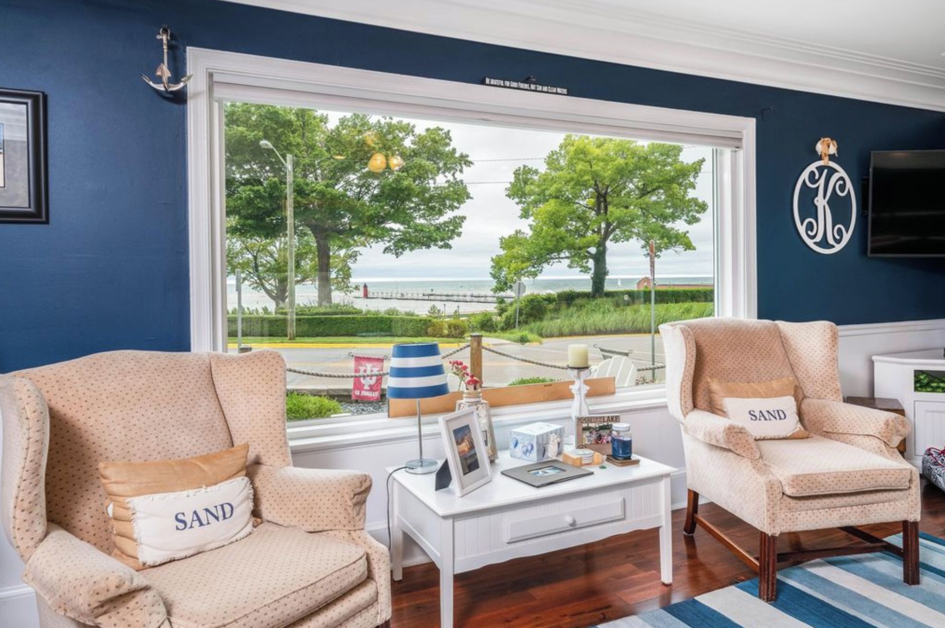 A room with two beige armchairs with 'SAND' pillows, a white side table with a picture frame, and a large window offering a view of the sea and trees.
