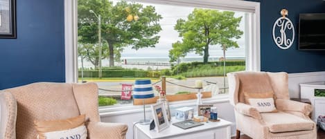 A room with two beige armchairs with 'SAND' pillows, a white side table with a picture frame, and a large window offering a view of the sea and trees.
