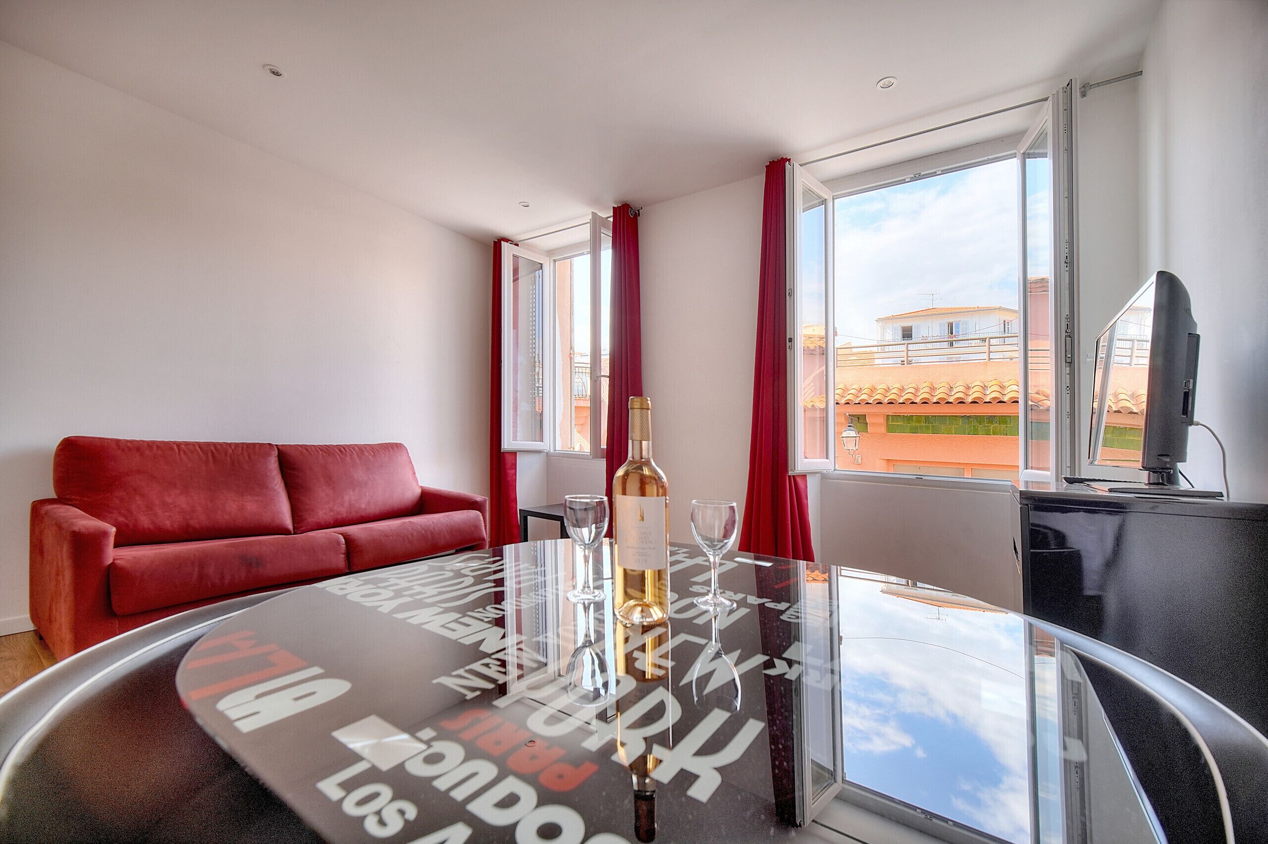 Une chambre d’hôtel avec un canapé rouge, une table en verre sur laquelle se trouvent une bouteille et des verres, et une vue sur les immeubles à travers de grandes fenêtres.