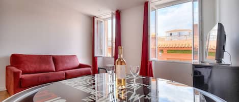 Habitación de hotel con un sofá rojo, una mesa de vidrio con una botella y vasos, y vistas a edificios a través de amplios ventanales.