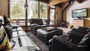 A cozy living room with leather sofas, a fireplace, a TV, and large windows overlooking a forest. - Alpine Manor Retreat Mountain Relaxation (Olympic Valley)