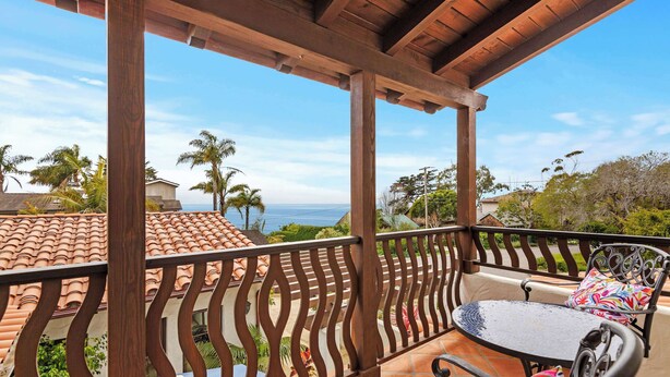 Un balcon extérieur abrité, doté d’une balustrade en bois, d’une table ronde avec un coussin, et offrant une vue sur l’océan et les palmiers.