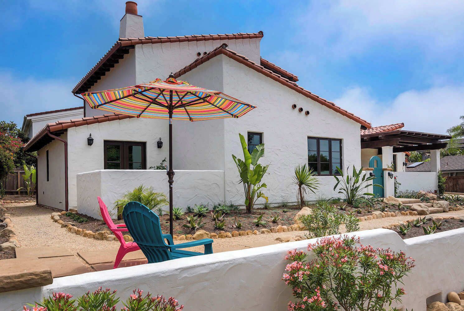 Une maison blanche avec un toit en tuiles, une terrasse agrémentée d’un parasol à rayures et un jardin où poussent différentes plantes et où l’on trouve des rochers.