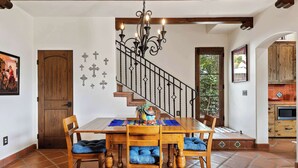 Un coin repas avec une table et des chaises en bois, un escalier doté d’une rampe en fer forgé, et une cuisine visible à travers une arche.