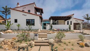 Une maison à deux étages, avec une façade blanche, un toit recouvert de tuiles rouges et un patio abrité. Des marches mènent à la porte d’entrée, et un jardin aménagé avec des rochers et des plantes se trouve à proximité.