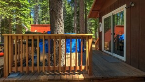 A wooden deck with a railing, a covered play area, and a building with a glass door. - Coziness Cabin - Cute Little Cabin in the Tahoe Woods! (South Lake Tahoe)