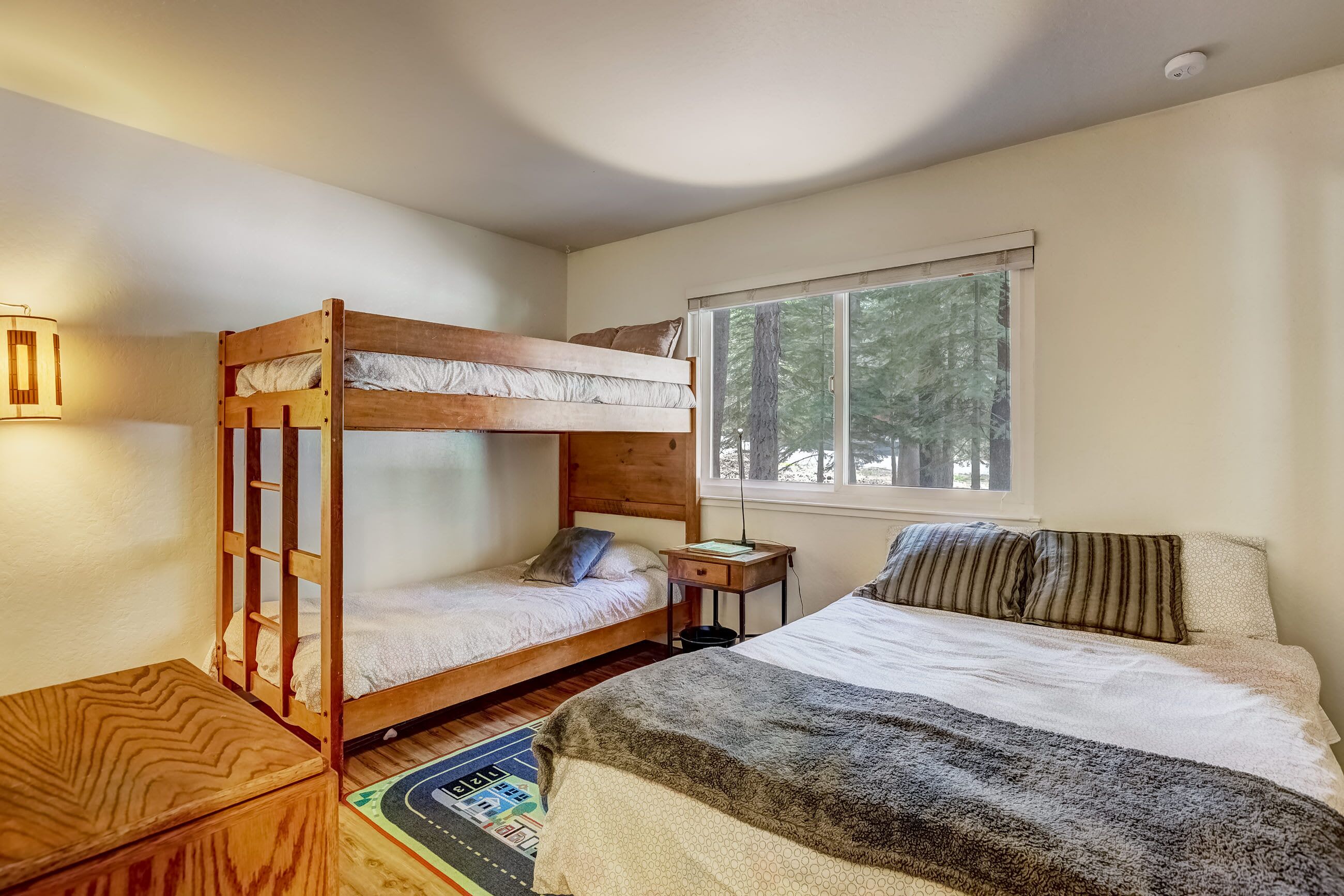 A room with a bunk bed, a single bed, a small wooden table, a nightstand, and a window with a view of trees.
