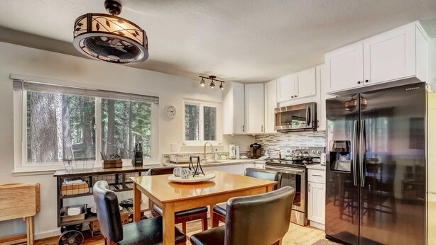 A modern kitchen with white cabinets, a stainless steel refrigerator, and a dining area with a wooden table and chairs. - Coziness Cabin - Cute Little Cabin in the Tahoe Woods! (South Lake Tahoe)