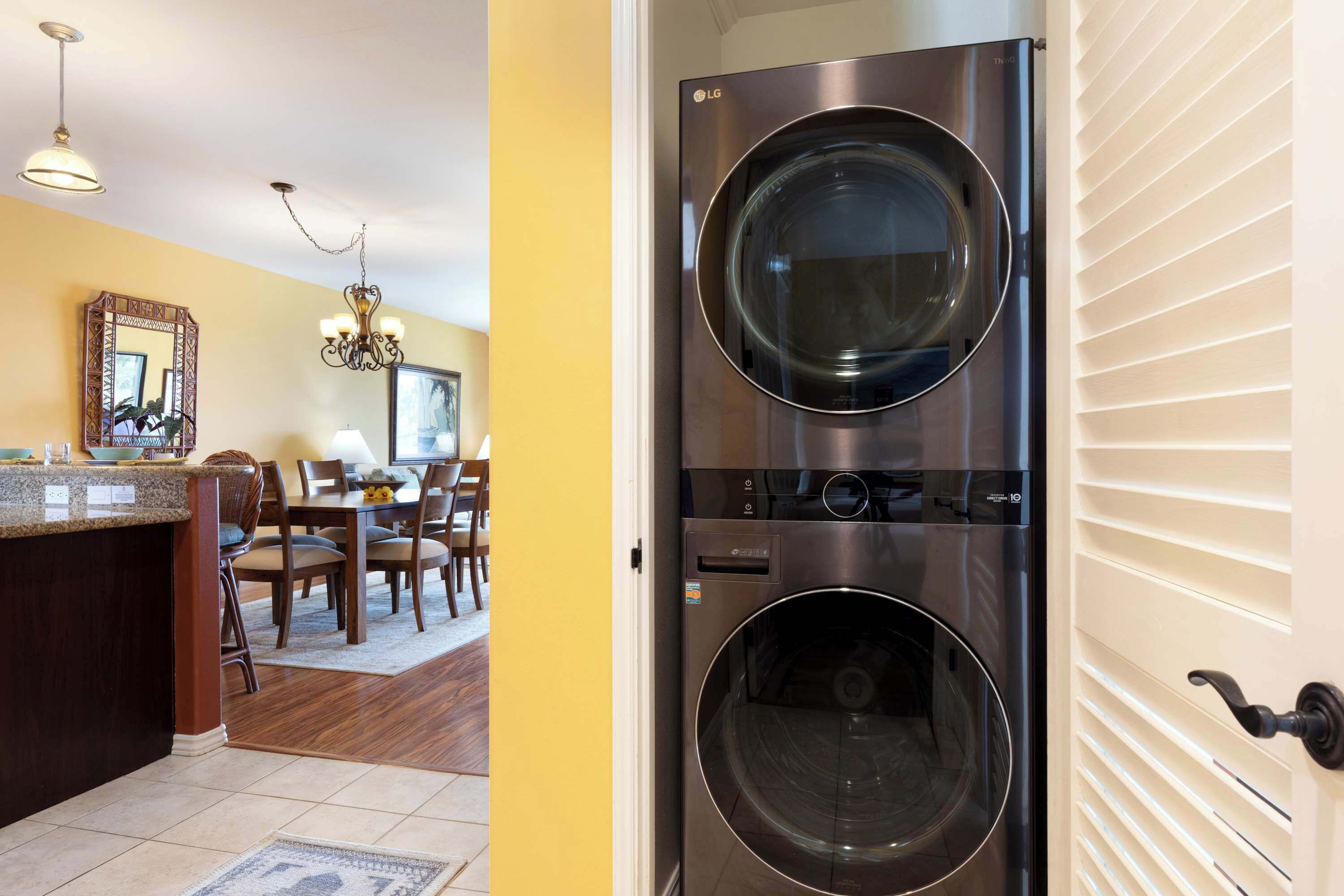 A laundry room with two stacked front-loading washing machines, a dining area with wooden chairs and a table, and a kitchen area with a granite countertop and a chandelier.