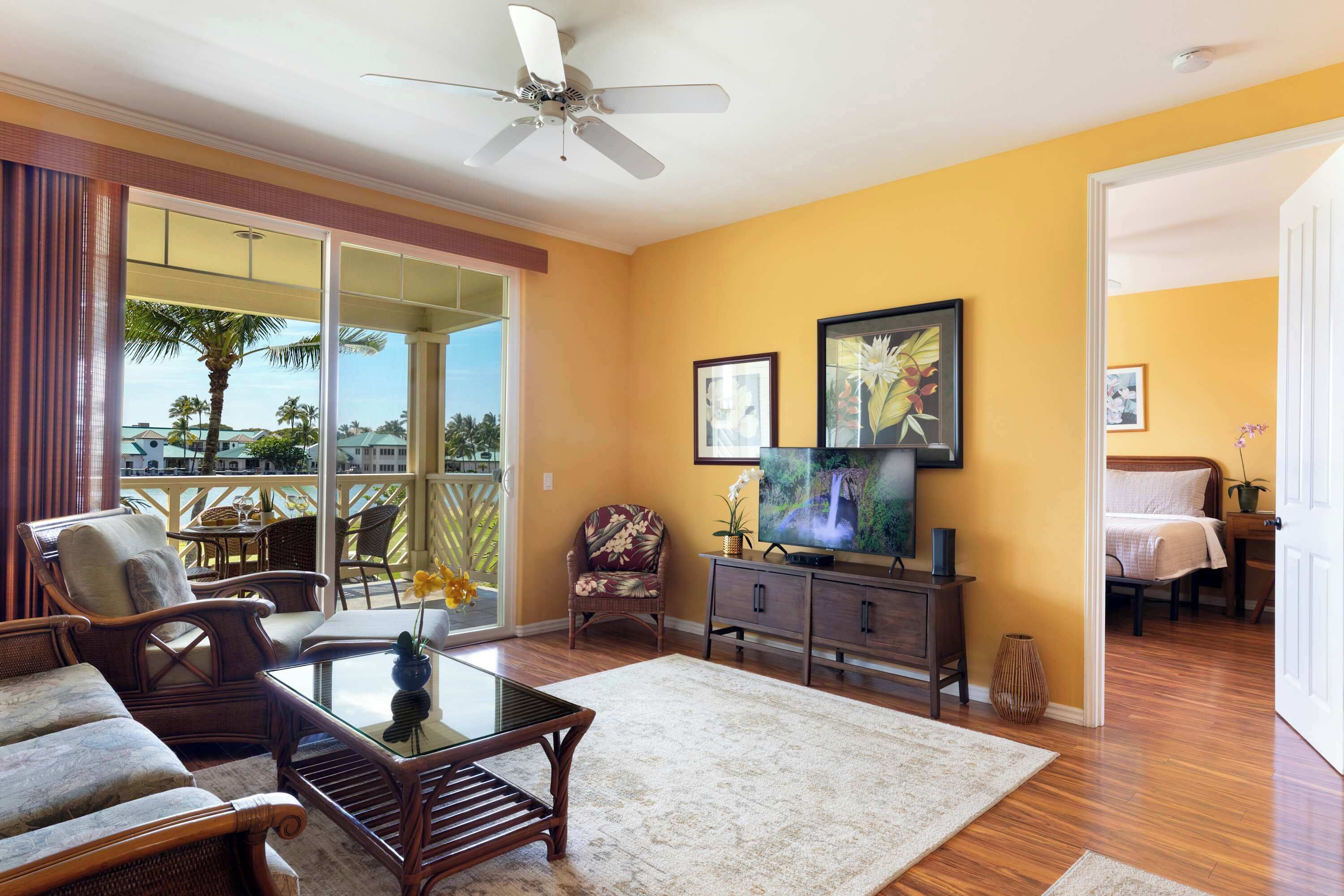 A living room with a sofa, armchairs, a coffee table, a TV, and a balcony with a view of palm trees.