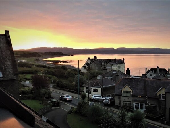 A coastal town with houses along the water, a road with cars, and a sunset in the background.
