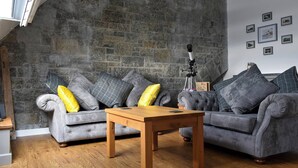 A cozy living room with a stone brick wall, two grey sofas with yellow cushions, a wooden coffee table, and a telescope on a tripod. - 2 bedroom accommodation in Criccieth (Criccieth)