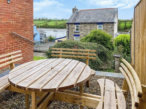 A wooden outdoor seating area with a table and benches, surrounded by pebbles and a hedge.