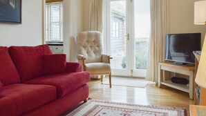 A living room with a red sofa, a white armchair, a television, and a large window with curtains. - 1 bedroom accommodation in Clopton, near Kettering (Clopton, near Kettering)