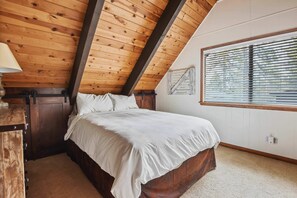 Ein Schlafzimmer mit Holzboden, einem Bett, einem Nachttisch, einem Fenster mit Jalousien und einer Wandleuchte.