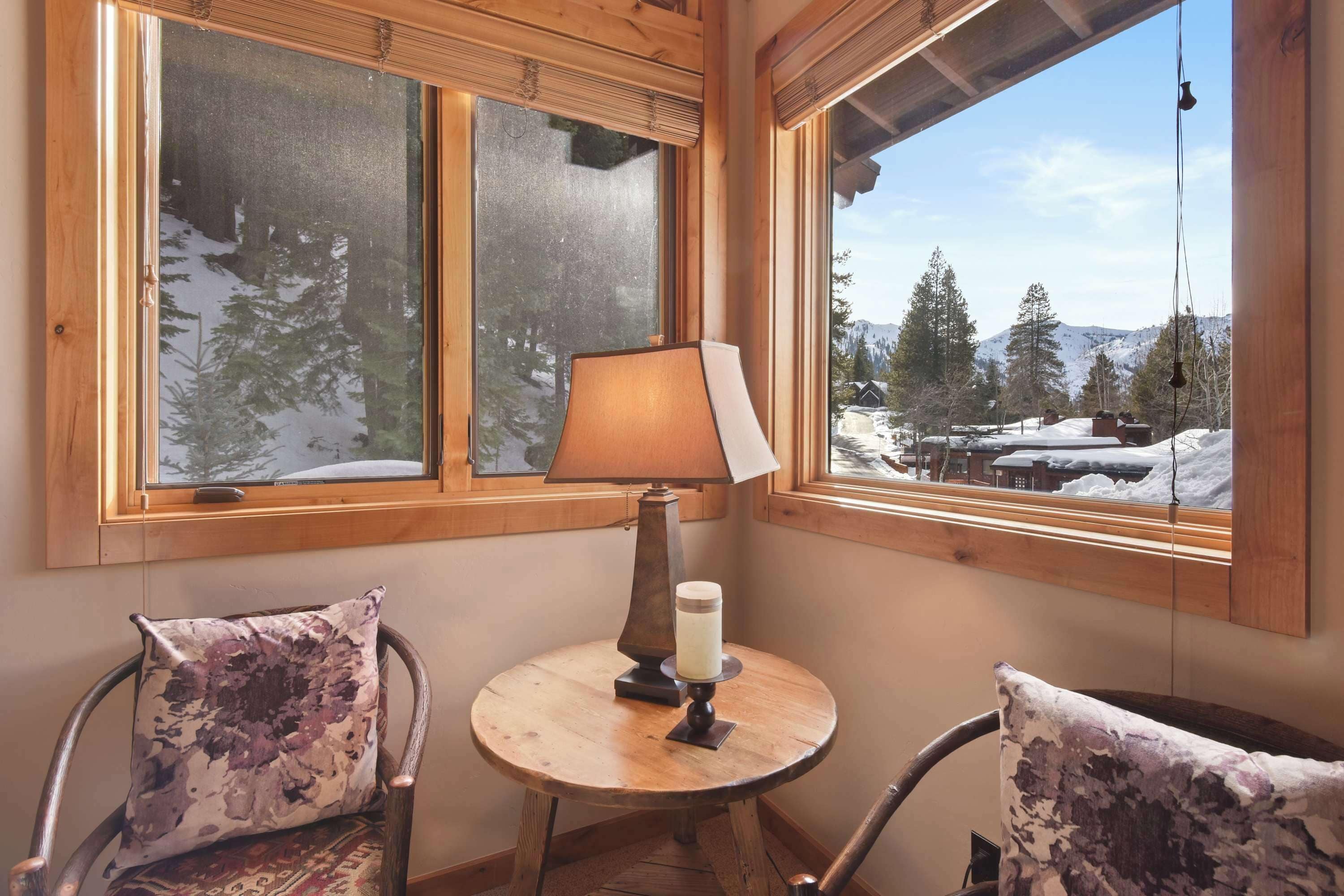 A cozy corner with a round wooden table, two chairs with patterned cushions, a lamp, and a candle. Large windows offer a view of a snowy landscape with trees and buildings.