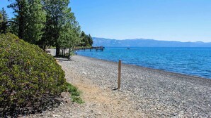 Una spiaggia di ciottoli con un molo di legno che si protende su un lago calmo, circondato da alberi e montagne sullo sfondo.