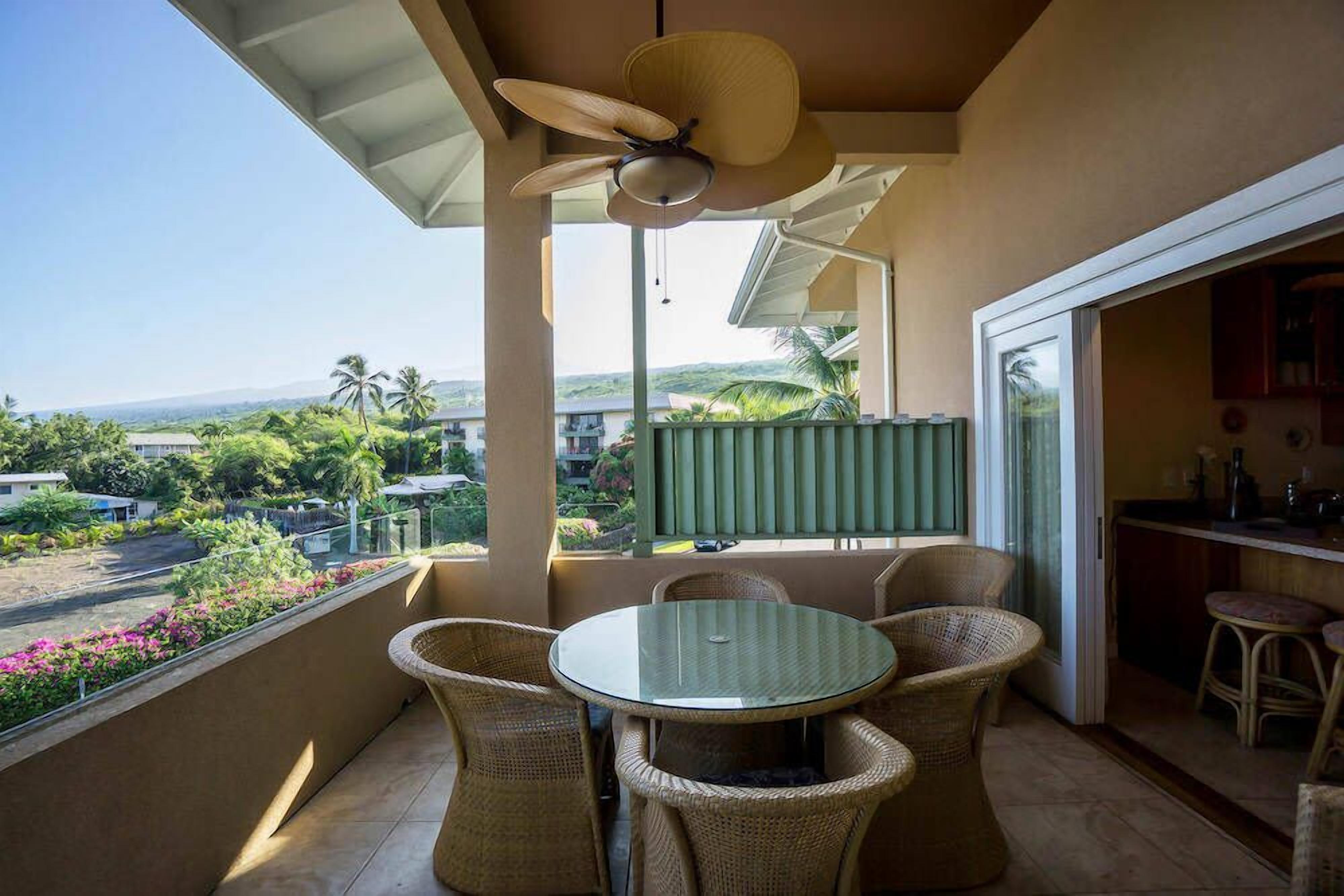 Un balcon doté d’une table ronde et de chaises en osier, offrant une vue imprenable sur un paysage luxuriant, avec des palmiers et l’océan à l’horizon.
