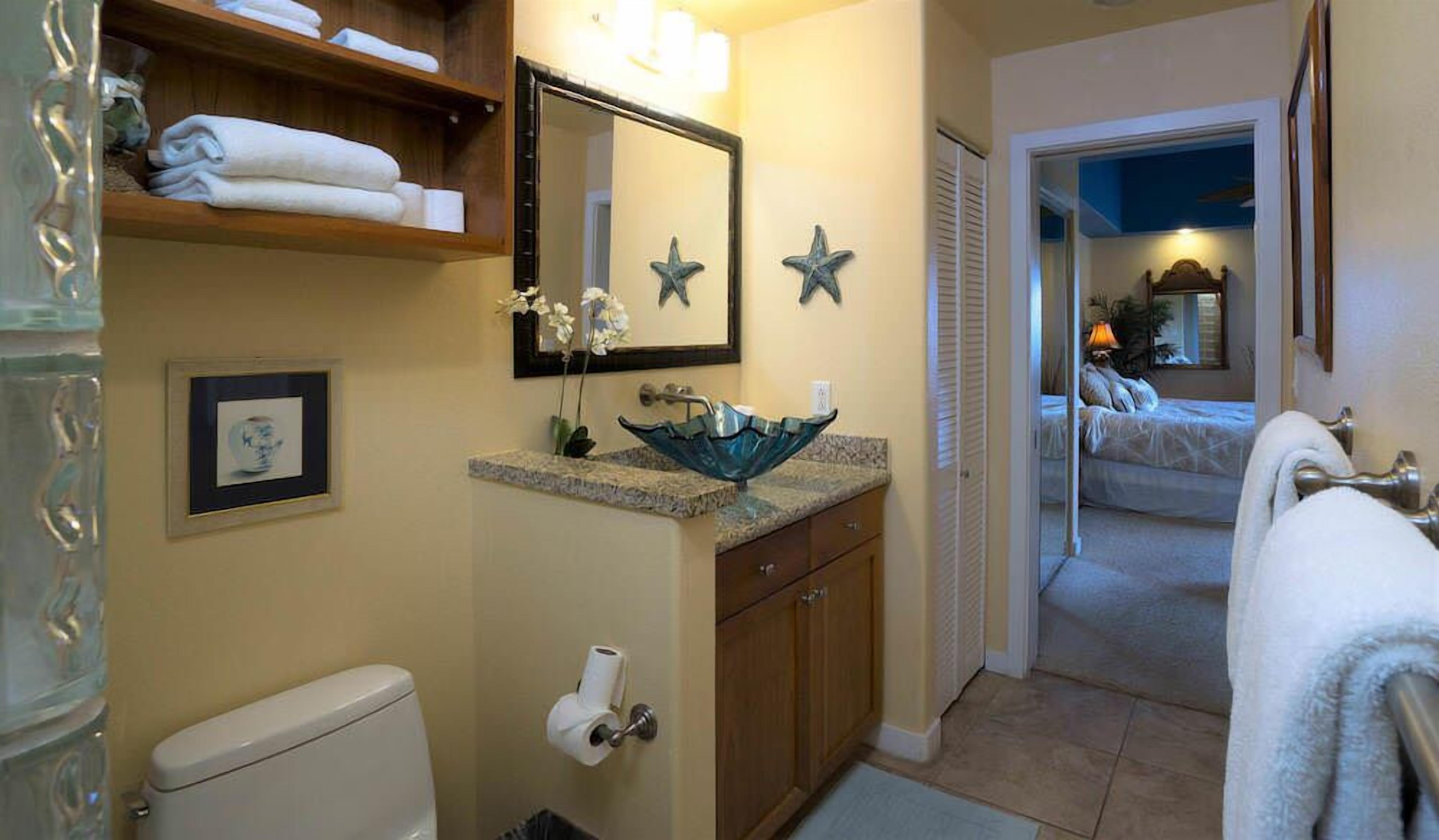 Baño con inodoro, lavamanos y zona de ducha. Se aprecia un estante con toallas y un cuadro en la pared.