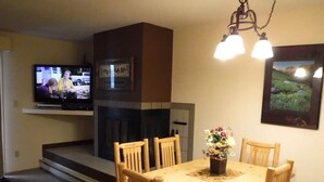 Un coin repas avec une table et des chaises en bois, une cheminée, un téléviseur fixé au mur et un tableau encadré au-dessus de la cheminée.