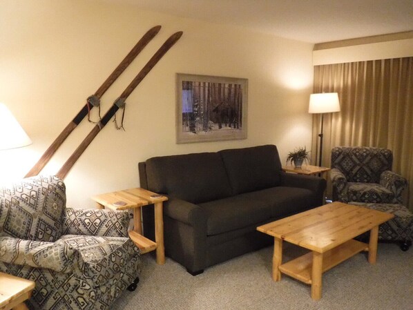 Een woonkamer met een bank, twee houten bijzettafeltjes, een fauteuil met een patroon, nog een fauteuil met een patroon, en een paar ski's die tegen de muur staan, plus een ingelijst kunstwerk.