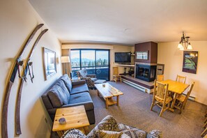A furnished living room with a sofa, coffee table, and dining table, a fireplace, a TV, and a large window with a mountain view. - Standard Condo w/ Slope Access (Whitefish)