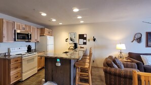 A compact kitchen with wooden cabinets, a microwave, a refrigerator, and a stove. There is a bar area with stools and a dining table. A couch and a lamp are visible in the living area. - Large, Split Level Condo (Whitefish)