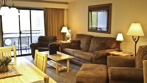 A cozy living room with a brown leather sofa, a wooden coffee table, and a dining table with chairs. There are two lamps on side tables and a large window with curtains. - Comfortable Condo Near Slopes (Whitefish)