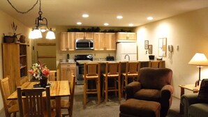A compact kitchen with wooden cabinets, a microwave, stove, and a refrigerator. There is a dining area with a table and chairs, and a sofa in the living area. - Nicely Furnished Condo with Valley Views (Whitefish)
