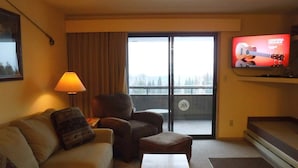 A hotel room with a sofa, armchair, ottoman, a television on the wall, and a balcony with a view. - Nicely Furnished Condo with Valley Views (Whitefish)