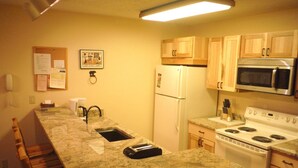 A kitchen with a granite countertop, white refrigerator, stainless steel microwave, and stove. There is a wooden bench along one wall and a corkboard with papers on the wall. - Charming Condo with Mountain Rustic Decor (Whitefish)