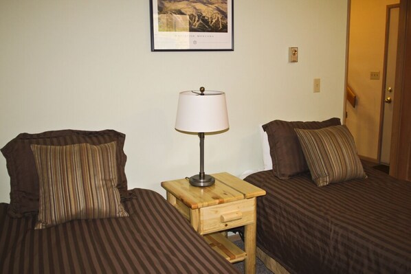 A room with two beds, each with striped bedding and pillows. There is a small wooden nightstand with a lamp between the beds. A framed picture hangs on the wall above the beds.