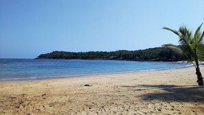 A beach with clear skies, calm water, and a small hill with vegetation in the background.