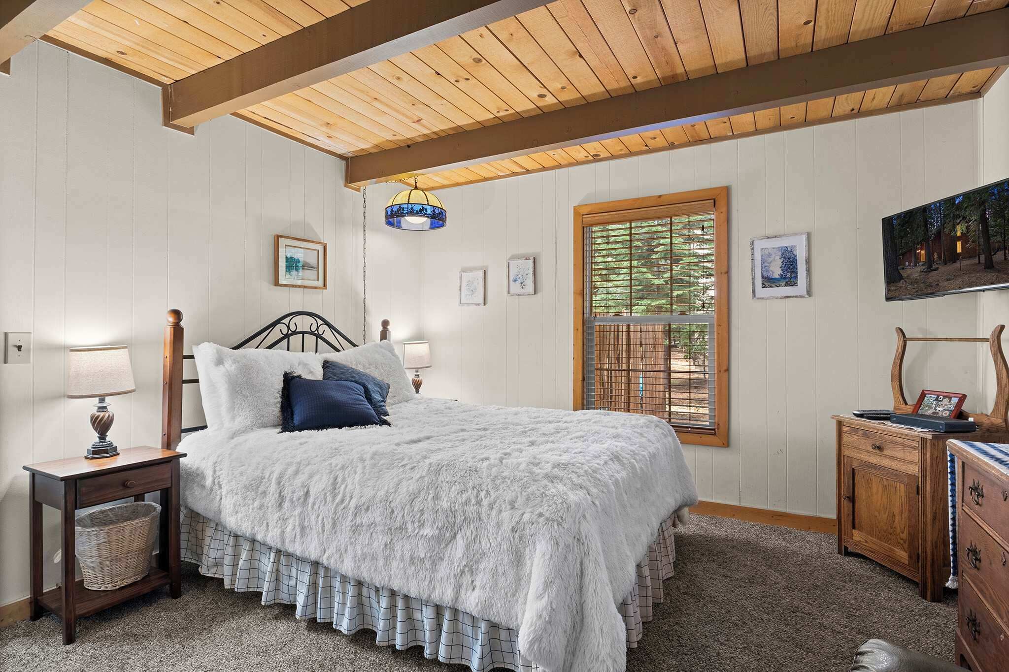 Un dormitorio acogedor con techo de madera, un ventanal con persianas, una cama con manta blanca mullida y una mesita de noche con lámpara.