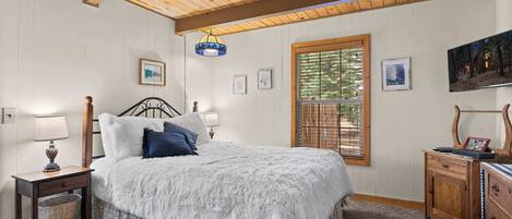 Un dormitorio acogedor con techo de madera, un ventanal con persianas, una cama con manta blanca mullida y una mesita de noche con lámpara.