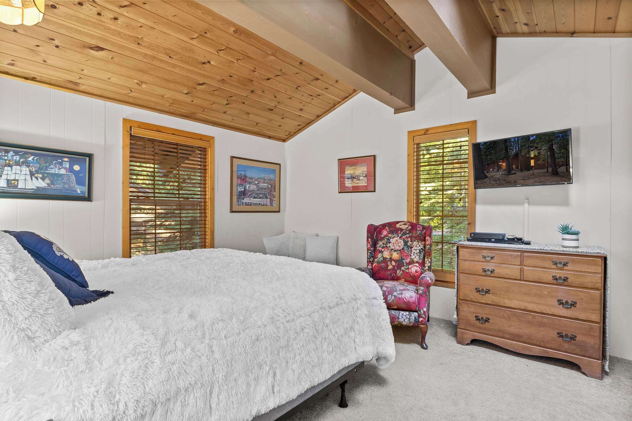 A bedroom with a bed, wooden ceiling, framed pictures, a dresser, and a television.