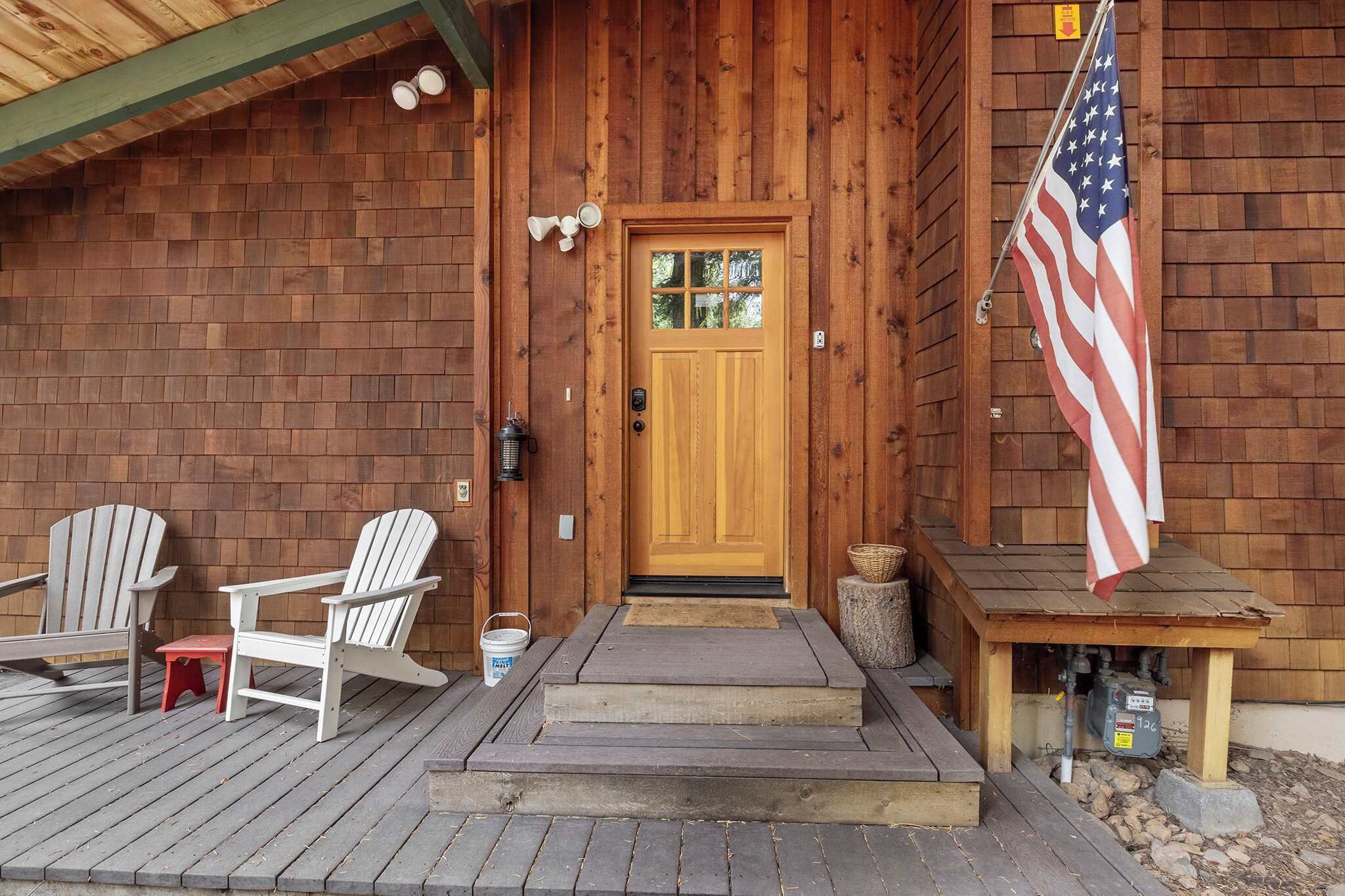 En stuga av trä med en ytterdörr, två Adirondack-stolar, ett litet bord och en amerikansk flagga.