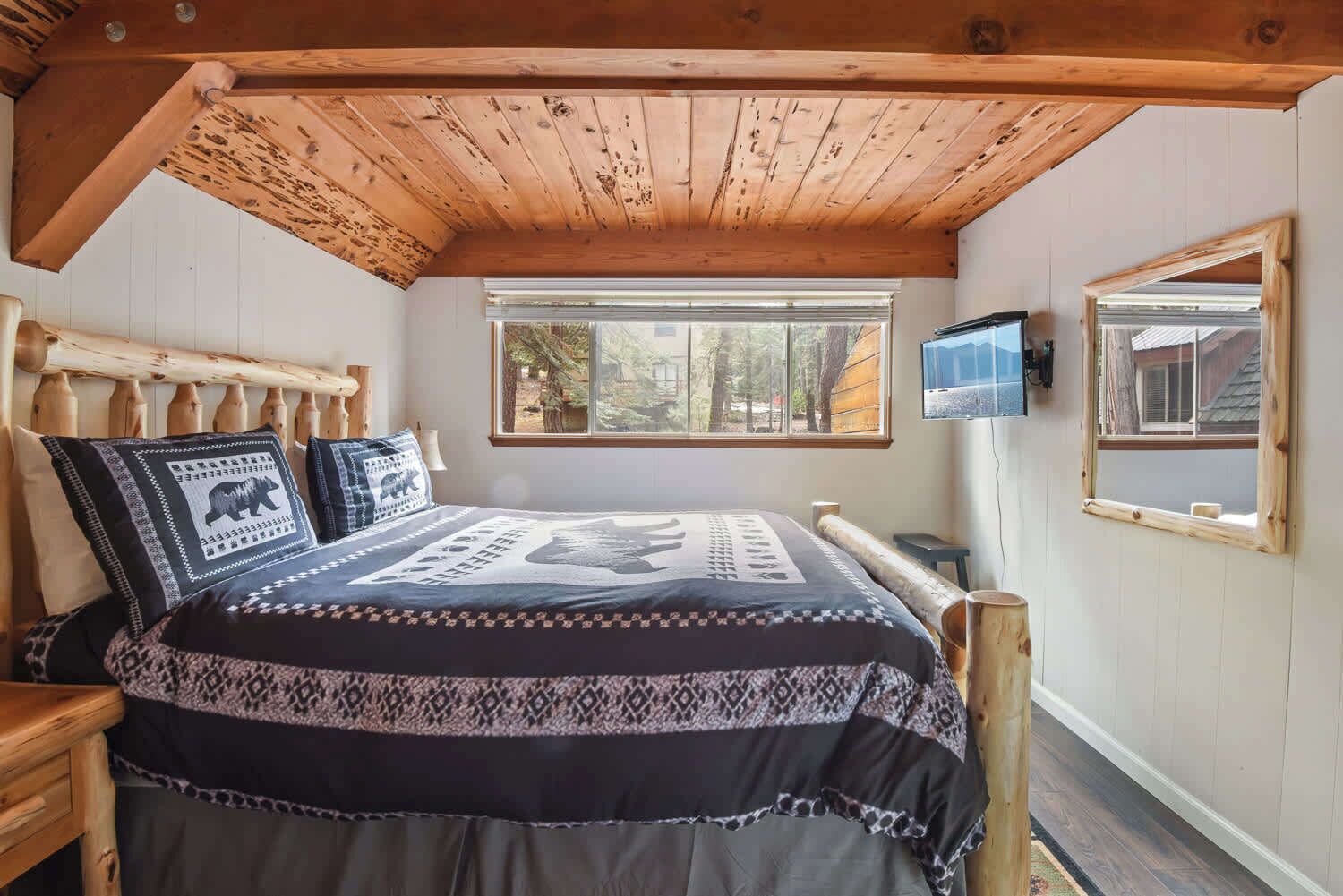 Une chambre avec un plafond en bois, une grande fenêtre, un lit avec une literie à motifs et un téléviseur fixé au mur.